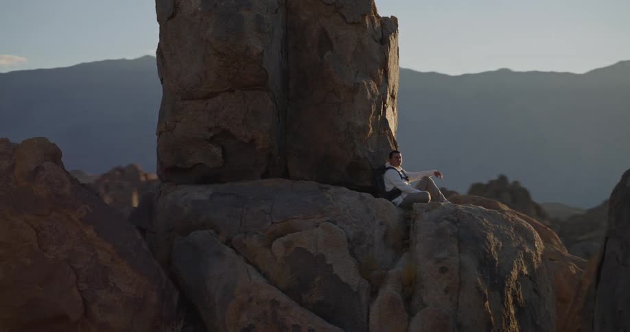 Man sitting on rock in desert landscape with vertical formation nearby and mountains in the background