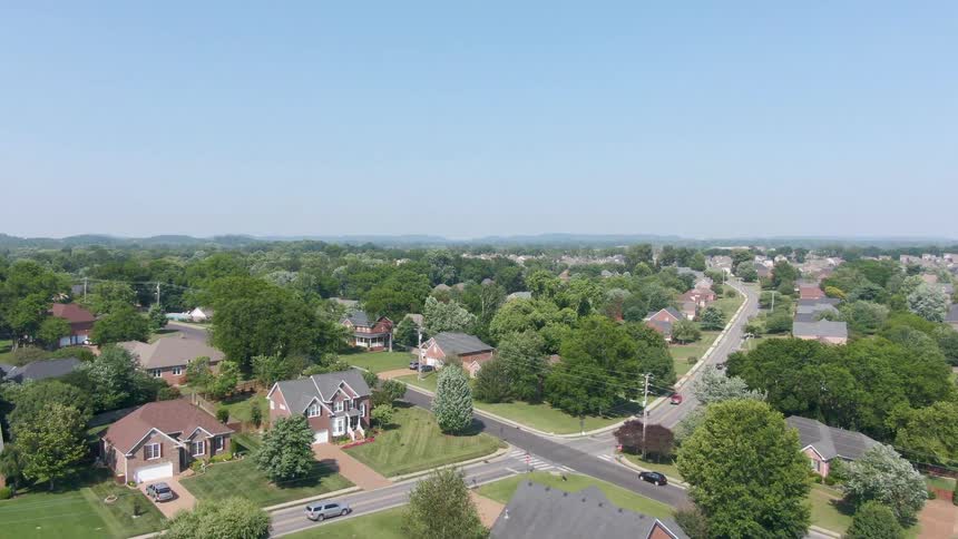An aerial view shows a sunny green suburban neighborhood with winding roads and neatly spaced houses