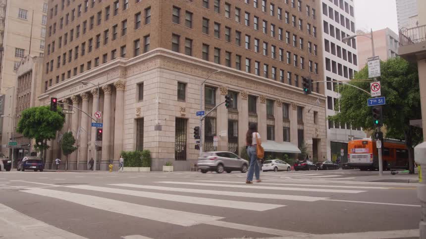A woman wearing a casual outfit jeans and a light top is walking from the right side of the frame toward the left across a marked crosswalkthe scene is dominated by a large classic stone bank building on the corner with traffic moving through the i