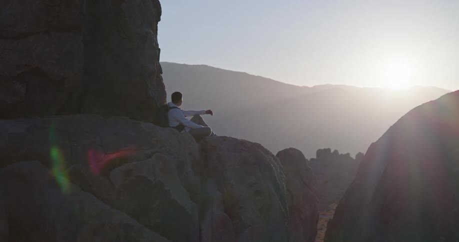 A man sitting on a large rock gazing out at a mountain range in the distance with the sun setting behind the mountains