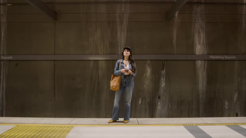 A young woman wearing a jean jacket jean trousers a hat and carrying a brown handbag stands at a train station terminal waiting for a train