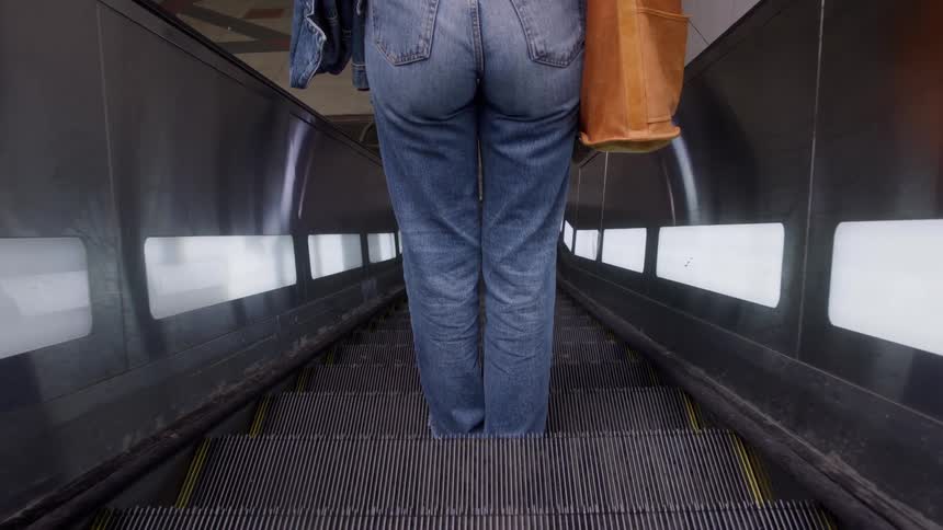 A woman rides the escalator down in a shopping mall at midday holding a brown handbag