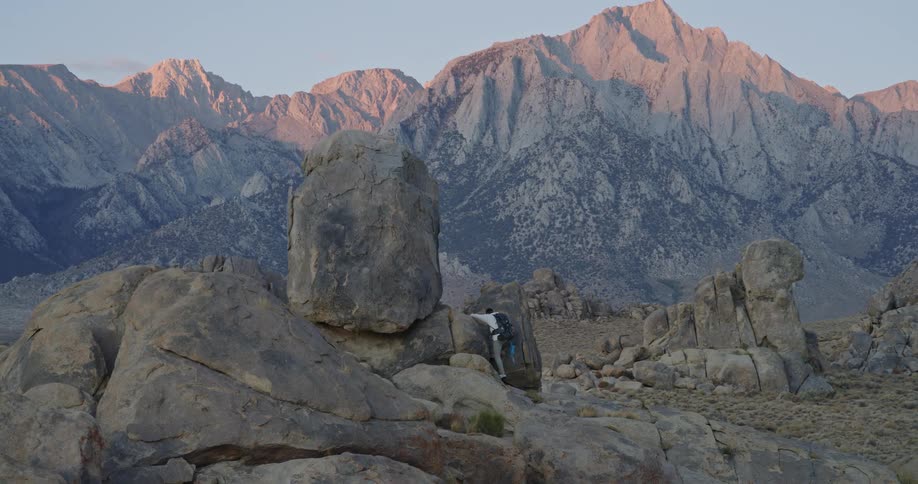Man carrying a backpack climbing into a tall rock on a wilderness area with a mountain range on the background outdoors during day time
