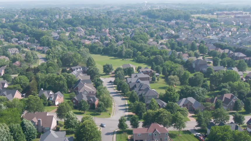 A sunny aerial view of a leafy suburban neighborhood shows tree lined streets cul de sacs and rows of tidy houses with wide green lawns