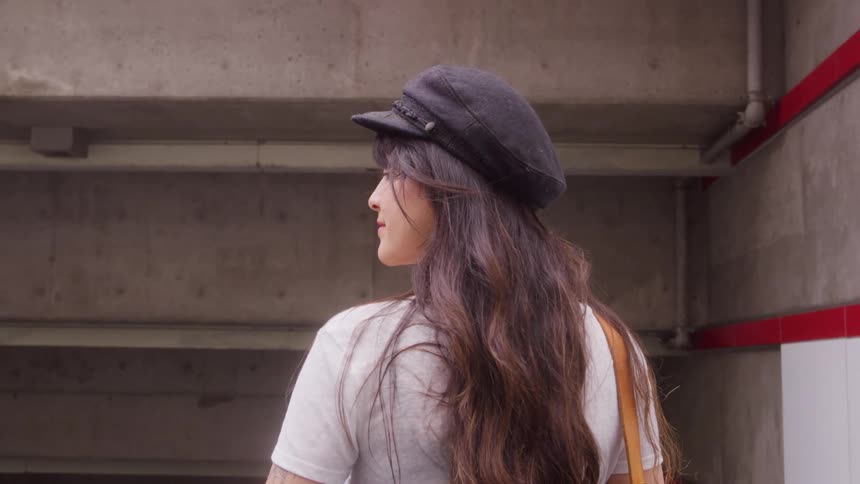 A woman with long brown hair and a black cap is seen from the back and side standing and looking upward within a structure made of gray concrete and white pipes with a red line running horizontally on the wall