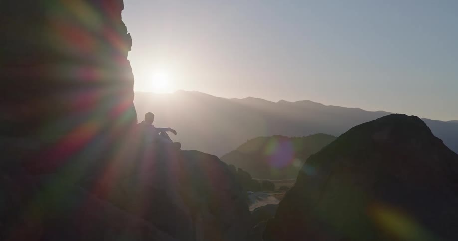 Man dressed in white sitting on rock with sun behind and mountains under clear sky