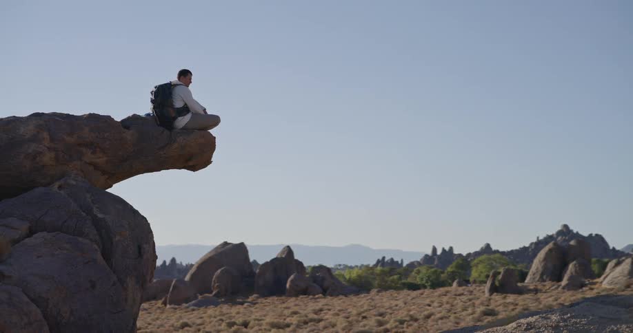 A man with a backpack sits on the end of a protruding rock that juts out over the ground looking out at a wide arid landscape of scattered rock formations and distant hills beneath a clear sky