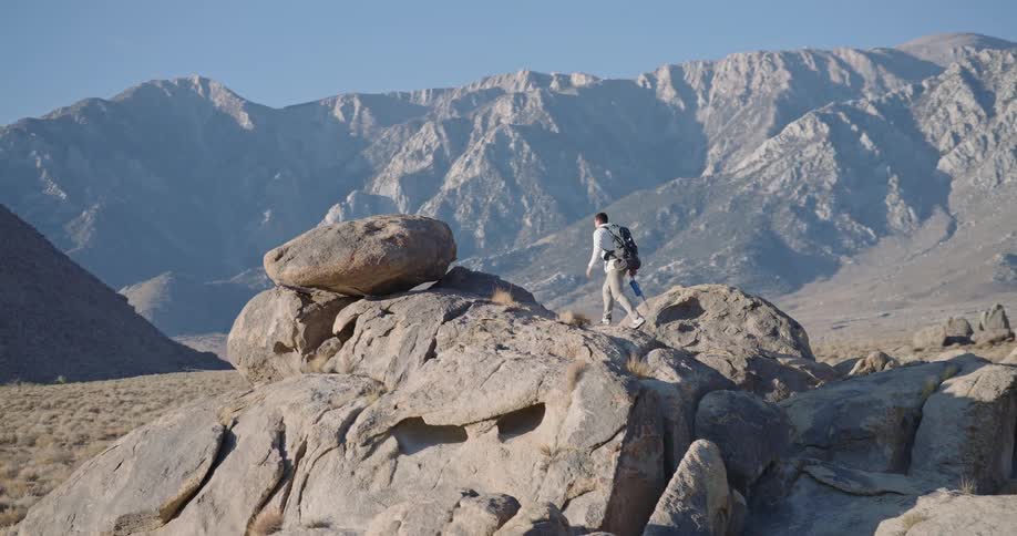 A lone hiker with a backpack carefully navigates a rocky terrain with a stunning mountain range in the background