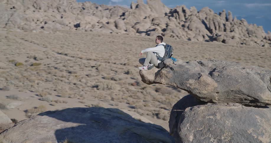 Man sitting on rock in desert landscape with sparse vegetation and mountains in the background under partly cloudy sky