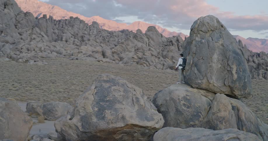 A young man hikes and climbs up rugged mountain terrain on a sunny day carrying a water bottle and a backpack