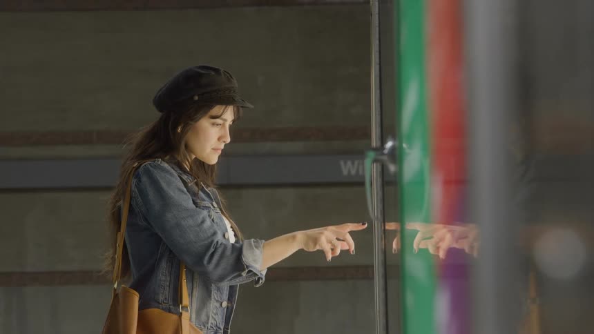 A young woman operates a computer screen while wearing a jean jacket and a hat carrying a brown handbag