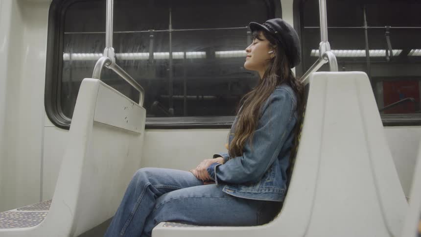 Woman sitting alone on side bench inside train car wearing denim jacket and black hat with contemplative gaze