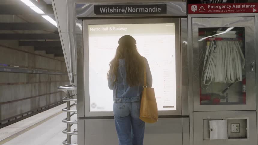 A young woman stands at a train station terminal waiting for a train holding a yellow handbag