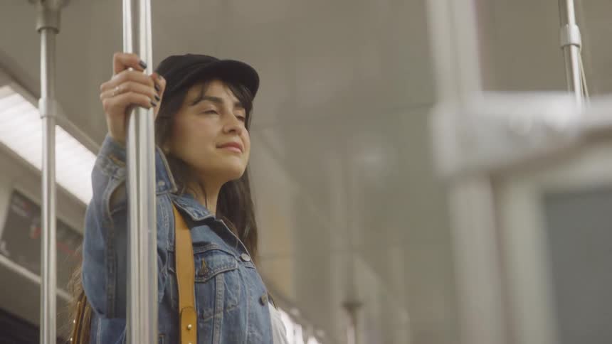 The video shows a mediumclose shot of a young woman with dark hair wearing a baseball cap and a denim jacket standing on a moving train or subway she is holding onto a silver overhead pole and looking out the windows to the right of the frame