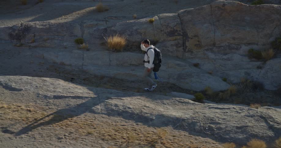 Man with black backpack walking through rocky terrain in arid environment wearing white shirt and jeans under soft early or late daylight