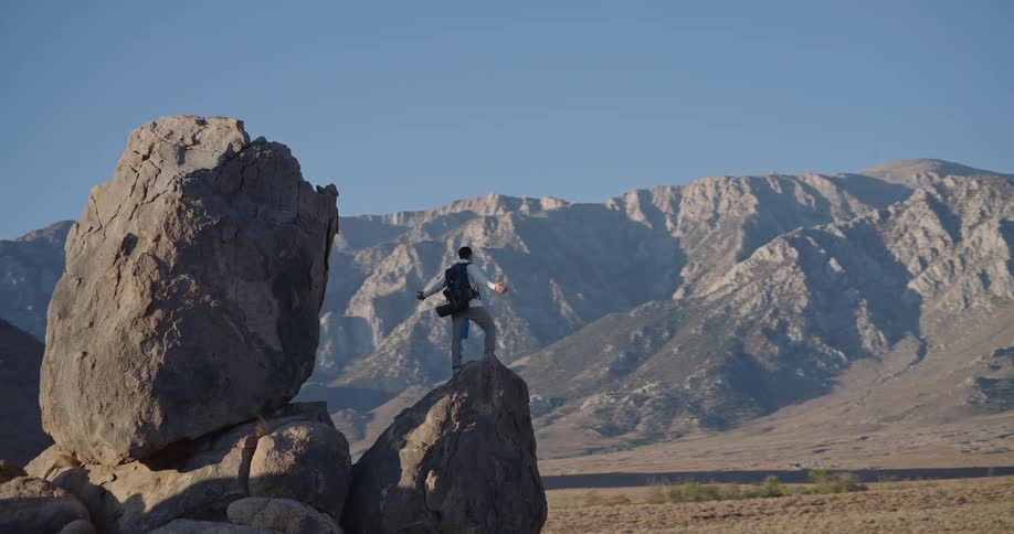 Person standing on sloped rock surrounded by mountains under clear sky