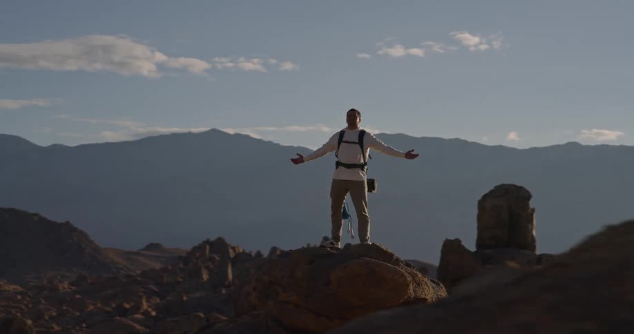 Person with arms outstretched on elevated rock facing the sun in mountainous landscape