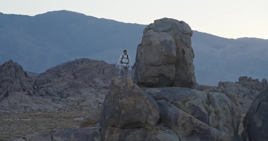 A male hiker stands calmly atop massive desert boulders at dusk gazing toward the pastel sky and distant ridgeline
