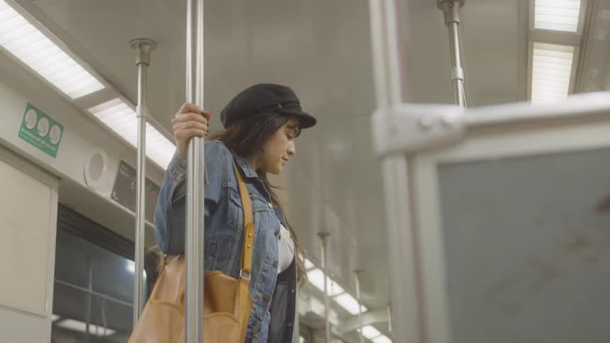 A young woman rides a train standing holding the pole wearing a hat and a jean jacket and carrying a brown handbag