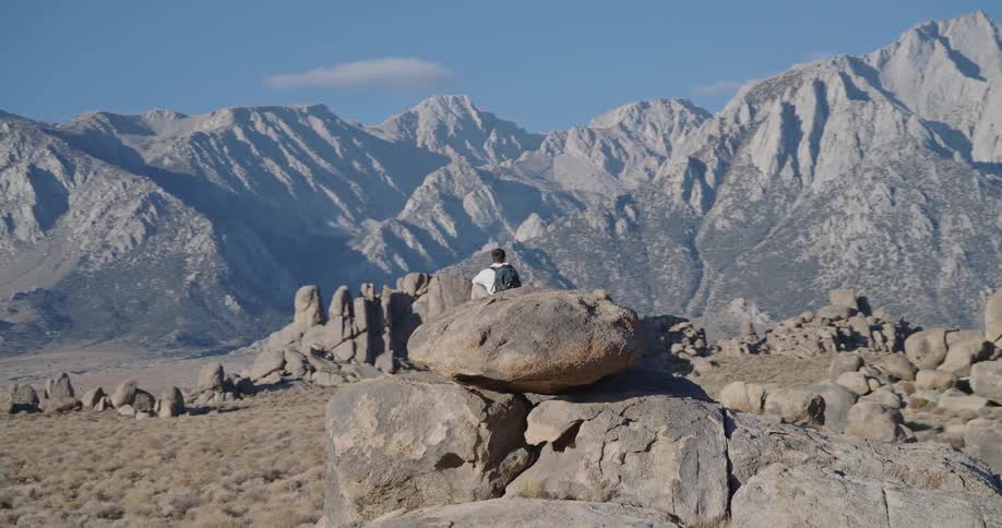 A hiker with a backpack sits on a large rock looking out at a vast majestic mountain range
