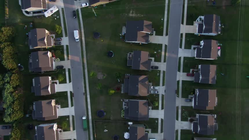 An overhead camera view looks directly down on a densely packed suburban residential neighborhood where rows of similar detached houses line a straight street grid with multiple vehicles and green lawns visible