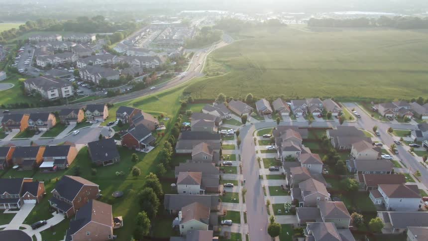 The video provides a high sweeping aerial shot of a modern suburban neighborhood backed by large open fields of green grass the camera is slowly moving forward over the densely packed houses showcasing the contrast between the residential development