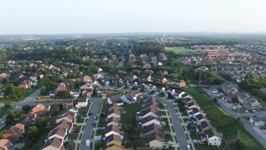 An aerial view slowly descends over a neatly planned suburban housing development showcasing rows of houses surrounded by lush dense green trees and forested areas the neighborhoods uniform layout and wide green buffers suggest a peaceful wellestabl