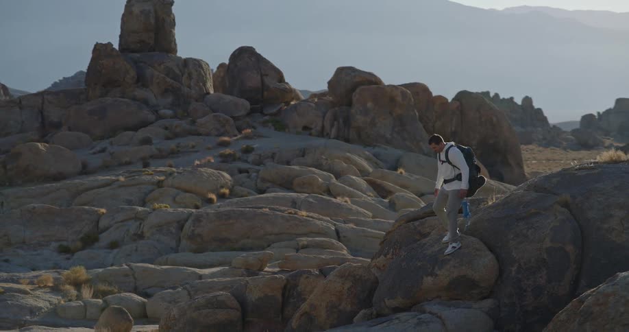 Man carrying a backpack walking through tall rocks surrounded with a mountain range in the background outdoors during day time