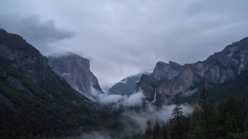 Granite cliffs shrouded in mist rise above forested valley under cloudy sky