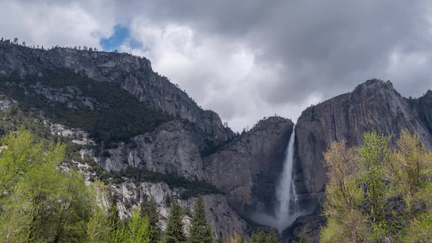 Waterfall cascades down a granite cliff under clouds with trees under cloudy sky