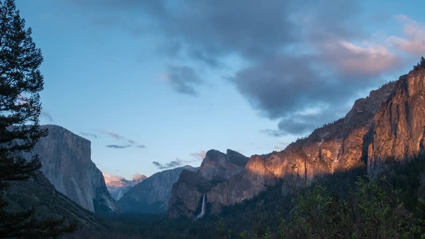 Rocky cliff rises behind forested valley under partly cloudy sky