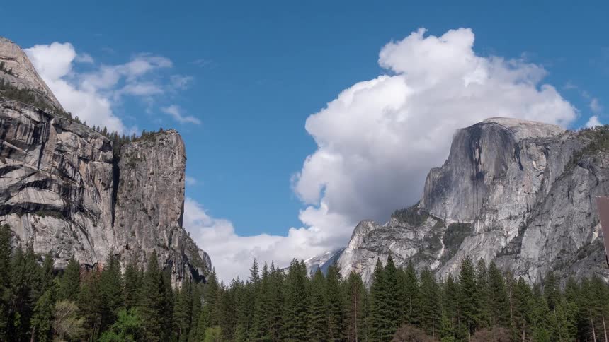 Granite cliffs rise among dense forest under blue sky with scattered clouds