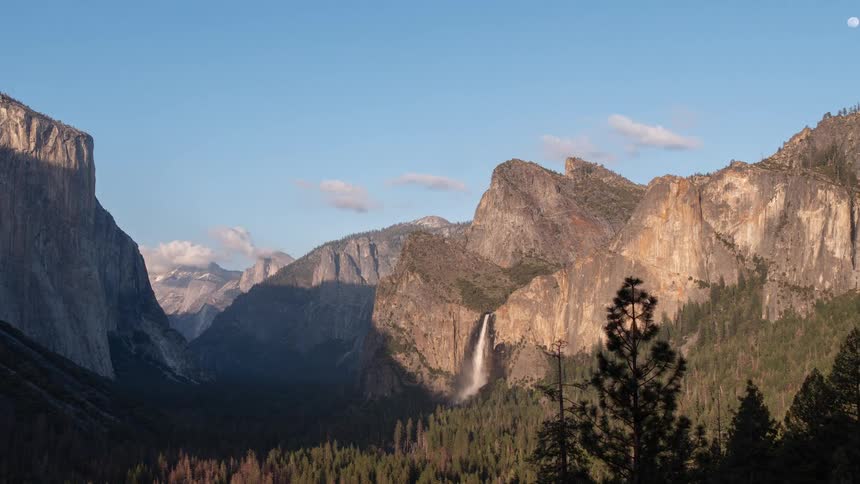 El capitan on the left and bridalveil fall in the center in forested valley under blue sky with clouds and moon