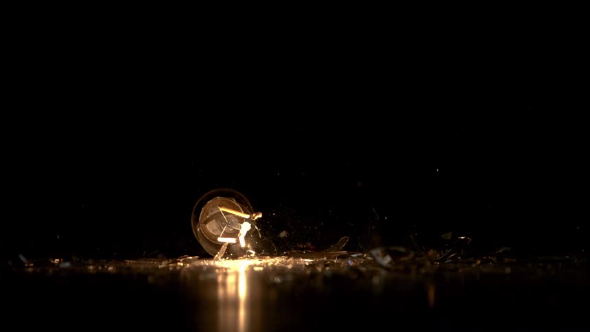 The video is a closeup shot of a filament light bulb sitting on a dark surface in a completely black room a hammer swings down and shatters the bulb in a slowmotion sequence accompanied by the bright flash of the filament breaking