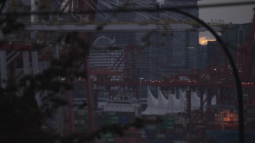 The view is dominated by numerous large dark red gantry cranes towering over stacks of shipping containers in the foregroundin the background a cluster of modern city skyscrapers is visible with a distinctive dome or rounded building structure sitti