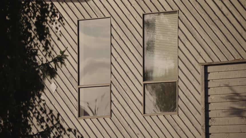The buildings siding consists of lightcolored horizontal wooden planks or similar material and there are two distinct tall narrow windows visible on the walltree shadow patterns are cast across the left side of the wall and one of the windows and