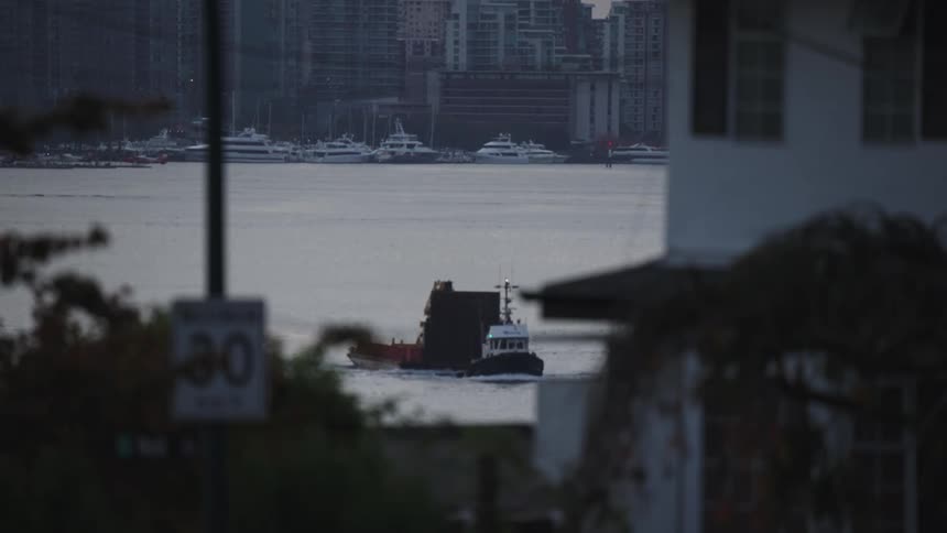 A small boat in the middle ground is pushing a large flat barge across a body of water toward the left surrounded by building structures and several docked yachts visible in the background
