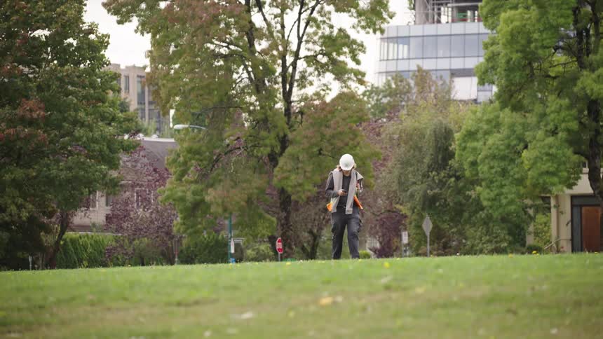 A construction worker walks across a grassy area near city buildings