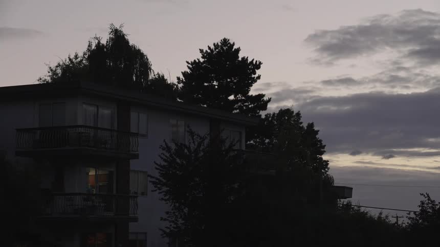 A quiet apartment building sits in silhouette at dusk one warm window glowing beneath an overcast sky and tall trees