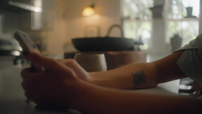 The video shows a closeup of a persons hands and arm which has a small tattoo as they scroll through content on a smartphone in a dimly lit kitchen the camera slowly pans up from the persons hands to their profile revealing a pensive expression as