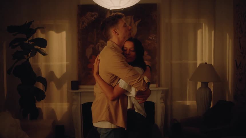 White young man and biracial young woman holding hands and hugging in a living room indoors during night time