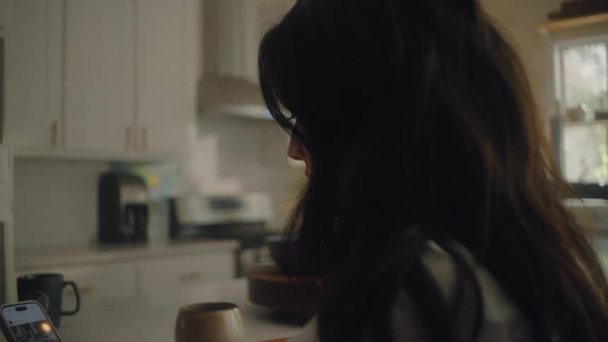 The back of the neck of a woman with brown hair who is in a kitchen using her cell phone