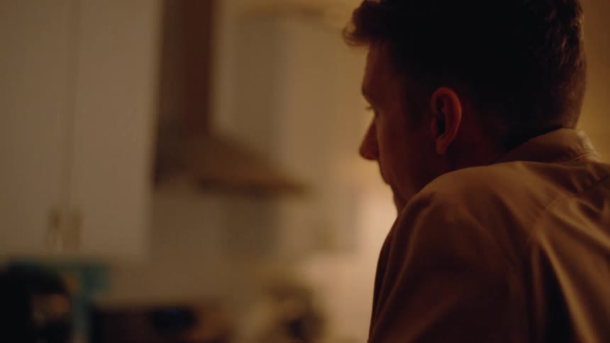 A woman sits in a kitchen talking to an unseen person with a serious expression before the camera focuses on the side profile of a man as they have a conversation the scene transitions to the couple standing in a warmly lit living room both appearing
