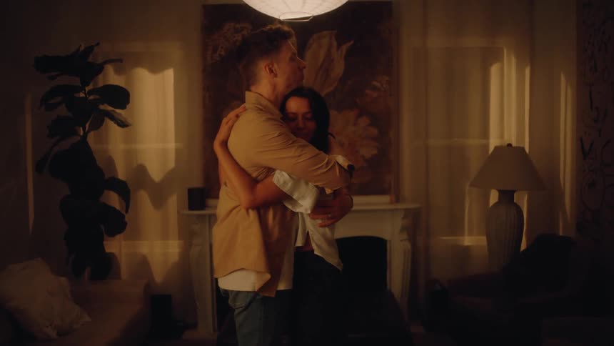 Young white man and young biracial woman holding hands and hugging in a living room indoors during night time