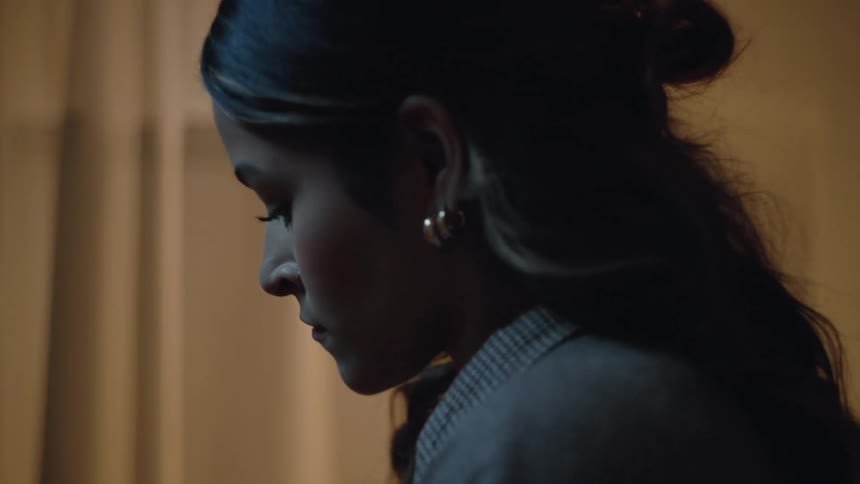 A close profile view video shows a young woman with dark hair pulled back and wearing a gray blazer looking down with a pensive expression in a dimly lit interior with vertical striped blinds or curtains in the background