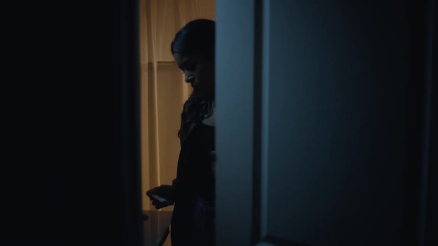 A young woman with long dark hair and a beanie is seen in dark silhouette briefly standing in a doorway while holding a bright phone before closing the door and plunging the view into darkness