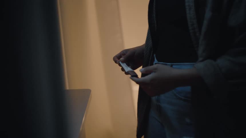 A person wearing blue jeans and a dark sweater or jacket stands in a dark room holding a white pregnancy test stick between their fingers with dim warm lighting highlighting their hands and the top of their thighs