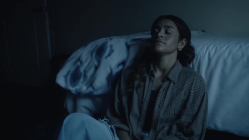 A young woman with dark hair wearing a plaid buttonup shirt and jeans sits on the floor of a dark bedroom closing her eyes and breathing deeply with a troubled expression before looking up and smiling briefly