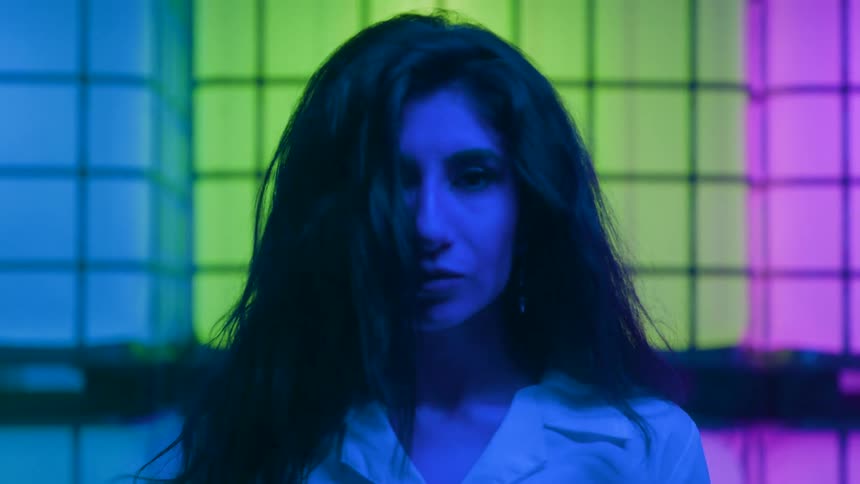 A woman lit in moody neon blues and greens stares downward her hair tousled and expression somber creating an eerie dramatic vibe
