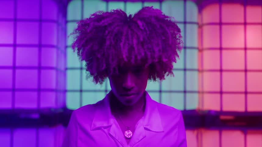 A person with curly purpletinted hair and a white shirt stands in front of a colorful backdrop made of stacked translucent containers illuminated by shifting neon lights the background transitions between bright purple green and orange squares givin